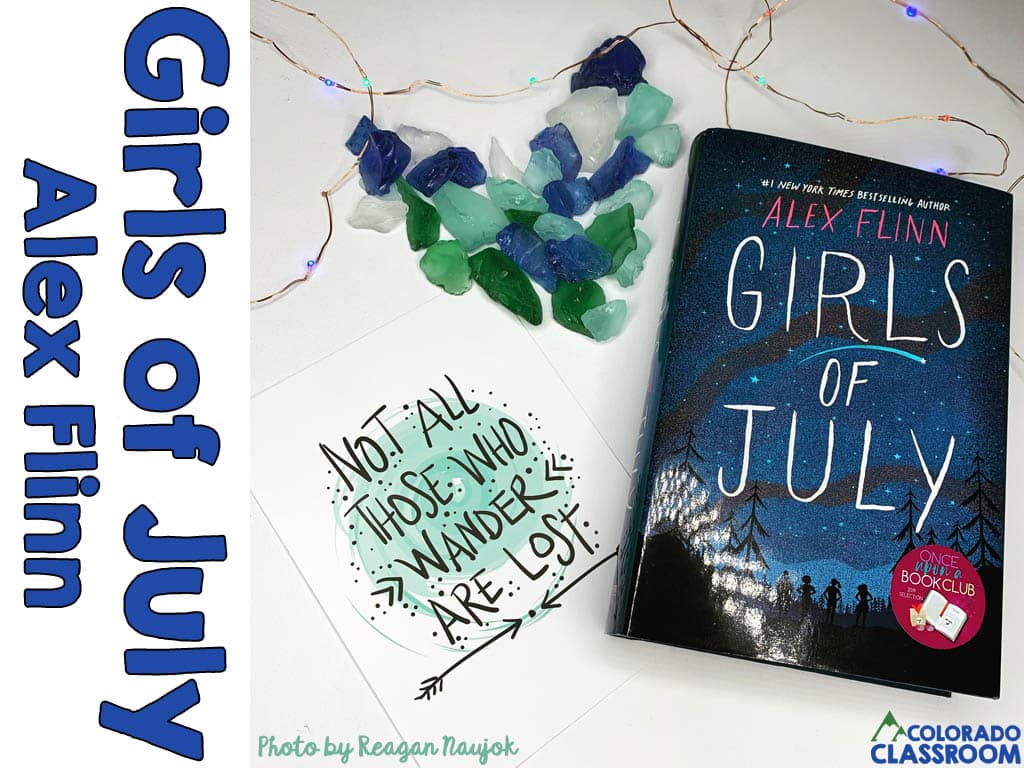 A book, Girls of July, in a stylized pose with a quote card, a heart made out of blue, green and white rocks, and some twinkling lights.