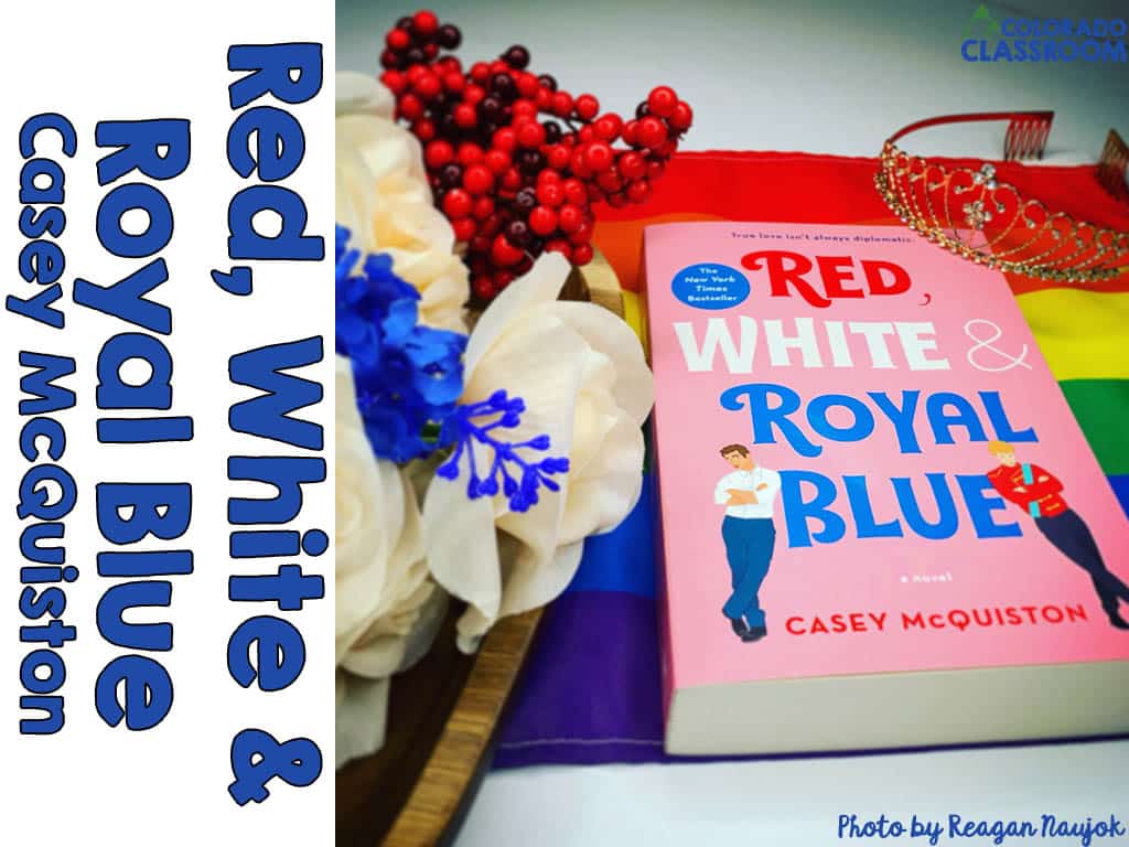 A book, Red White & Royal Blue, in a stylized pose with some white and blue silk flowers, plastic red berries, a wooden tray, a Pride flag, and a tiara.